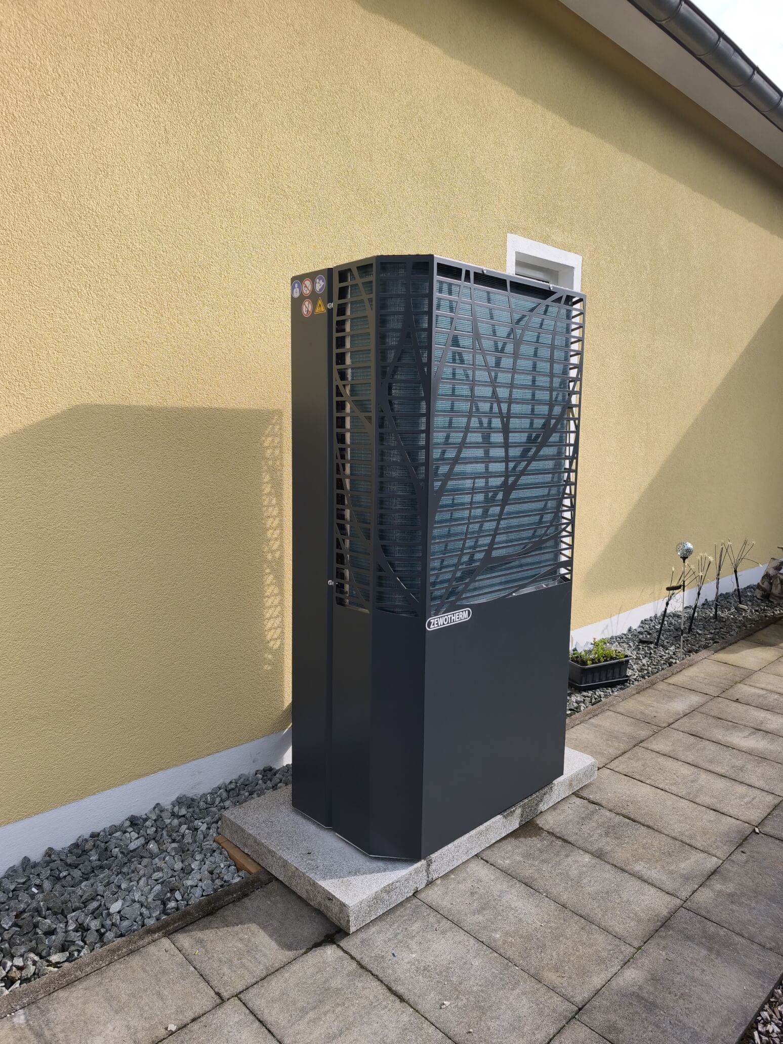 Effiziente Wärmepumpen-Heiztechnik in einem modernen Außenbereich Ein moderne Wärmepumpe steht auf einem grauen Steinboden vor einer gelben Wand, umgeben von Kies und Pflanzen.