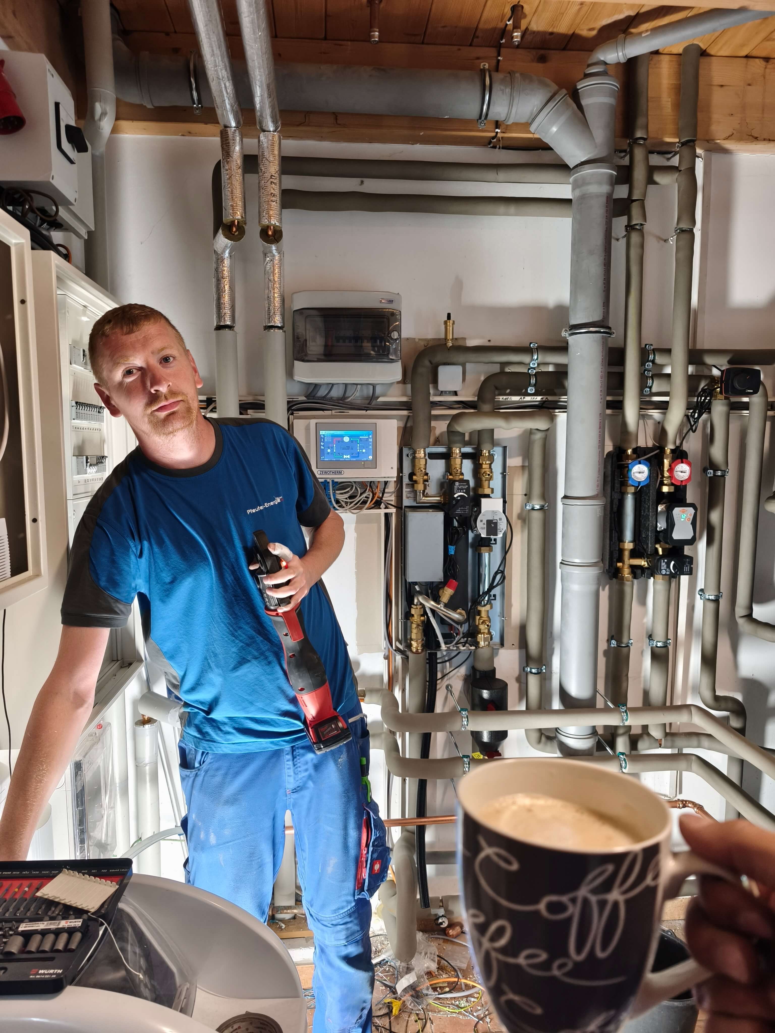 Moderne Heizungsinstallation mit einem Pfeufer-Energie Fachmann im Einsatz Ein Pfeufer-Energie Monteur in blauer Kleidung arbeitet an der Installation der Wärmepumpe in einem Technikraum, während er ein Werkzeug in der Hand hält.